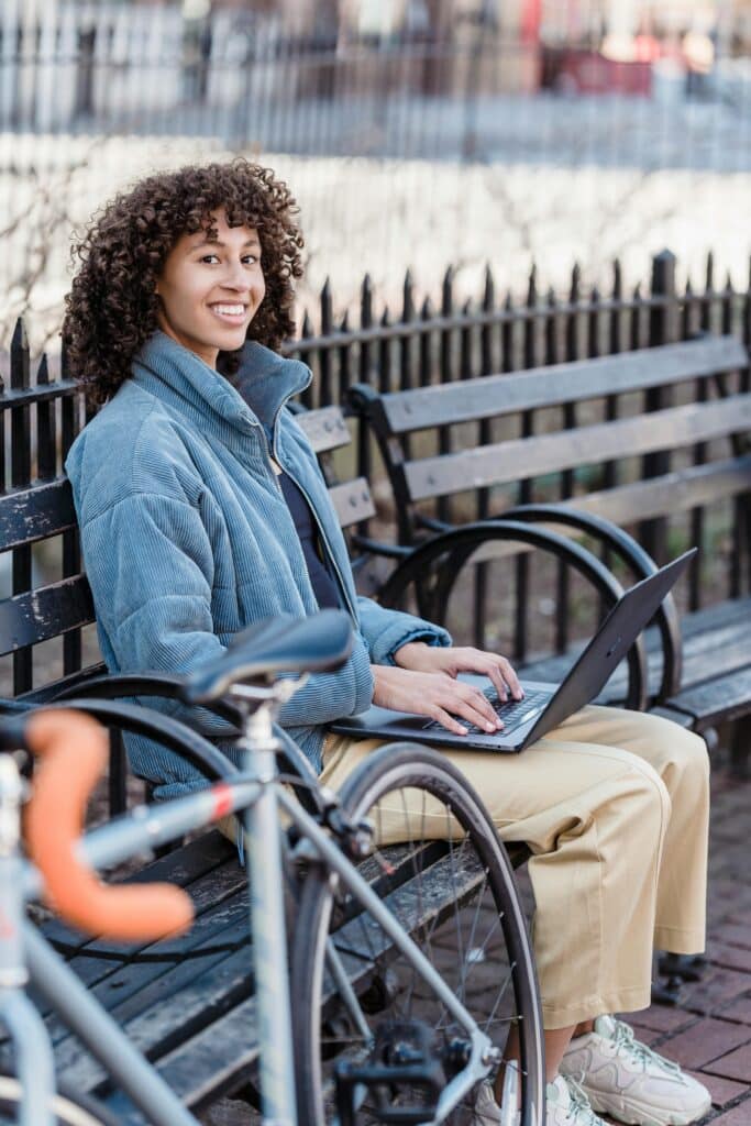 A smiling woman sitting on a park bench, working on a laptop, with a bicycle parked in the foreground and a metal fence and trees in the background.